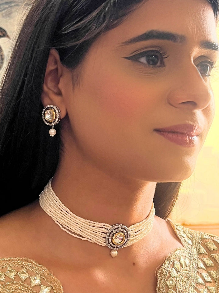 A woman wears the Pearl White Renu Necklace Set, featuring a multi-strand white pearl choker with Kundan and White accents, a central jeweled pendant, and matching round earrings adorned with dangling pearls. Kundan choker necklace accentuates her look.