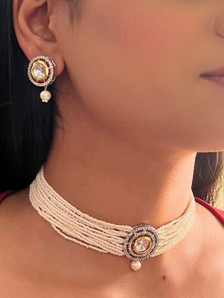 A woman wears the Pearl White Renu Necklace Set, featuring a multi-strand white pearl choker with Kundan and White accents, a central jeweled pendant, and matching round earrings adorned with dangling pearls. Kundan choker necklace accentuates her look.