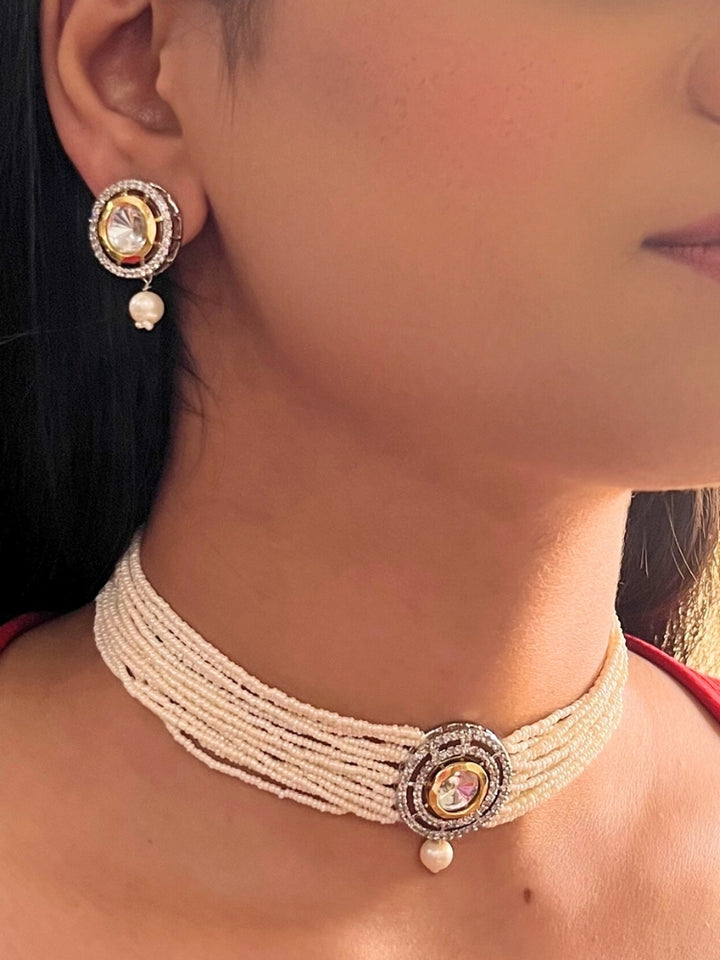 A woman wears the Pearl White Renu Necklace Set, featuring a multi-strand white pearl choker with Kundan and White accents, a central jeweled pendant, and matching round earrings adorned with dangling pearls. Kundan choker necklace accentuates her look.