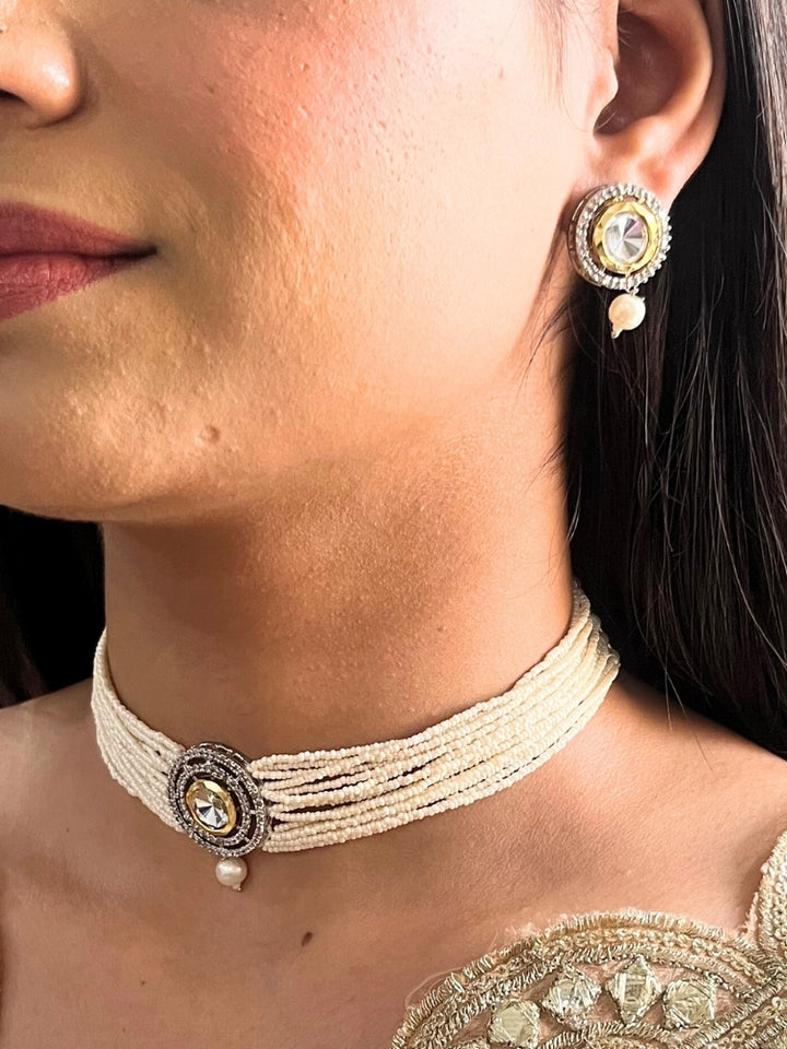A woman wears the Pearl White Renu Necklace Set, featuring a multi-strand white pearl choker with Kundan and White accents, a central jeweled pendant, and matching round earrings adorned with dangling pearls. Kundan choker necklace accentuates her look.