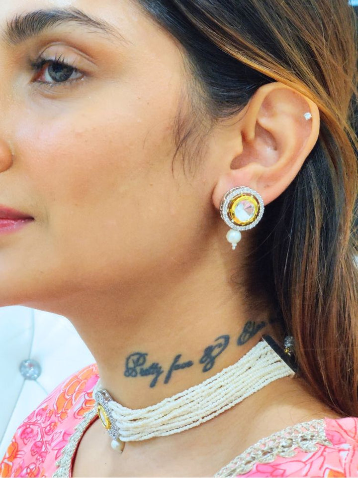 A girl looking on the side wearing The Pearl White Renu Necklace Set features a beaded choker with a bold circular pendant and matching stud earrings, embellished with gold, silver, American diamonds, and pearl accents. 