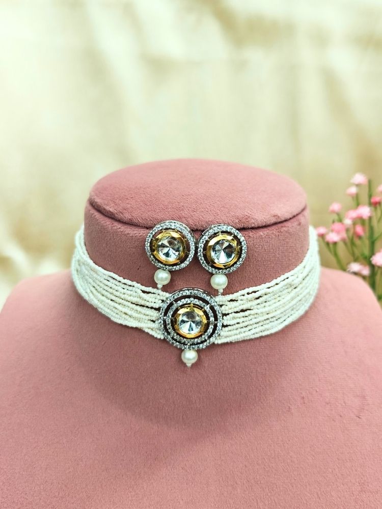 The Pearl White Renu Necklace Set features a beaded choker with a bold circular pendant and matching stud earrings, embellished with gold, silver, American diamonds, and pearl accents. Displayed on a pink stand with pink flowers in the background.