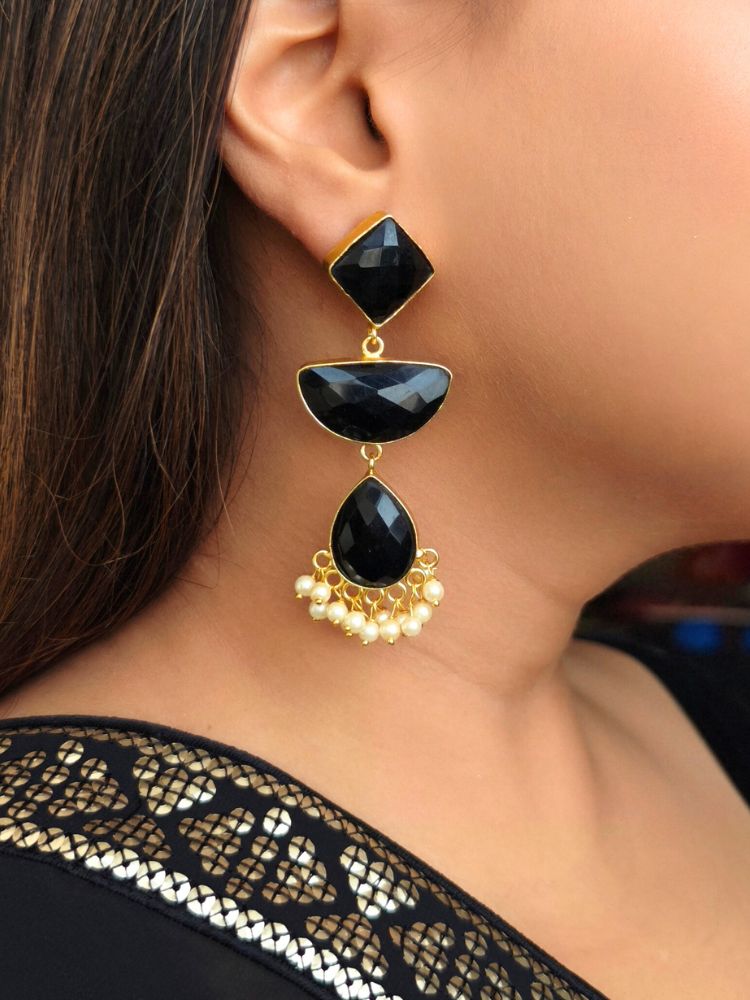 A close-up of a womans ear wearing Penelope Black Earrings—large, gold-framed black earrings with Monalisa stones and white pearl accents—paired with a gold-embroidered black outfit, perfect for a wedding ceremony.