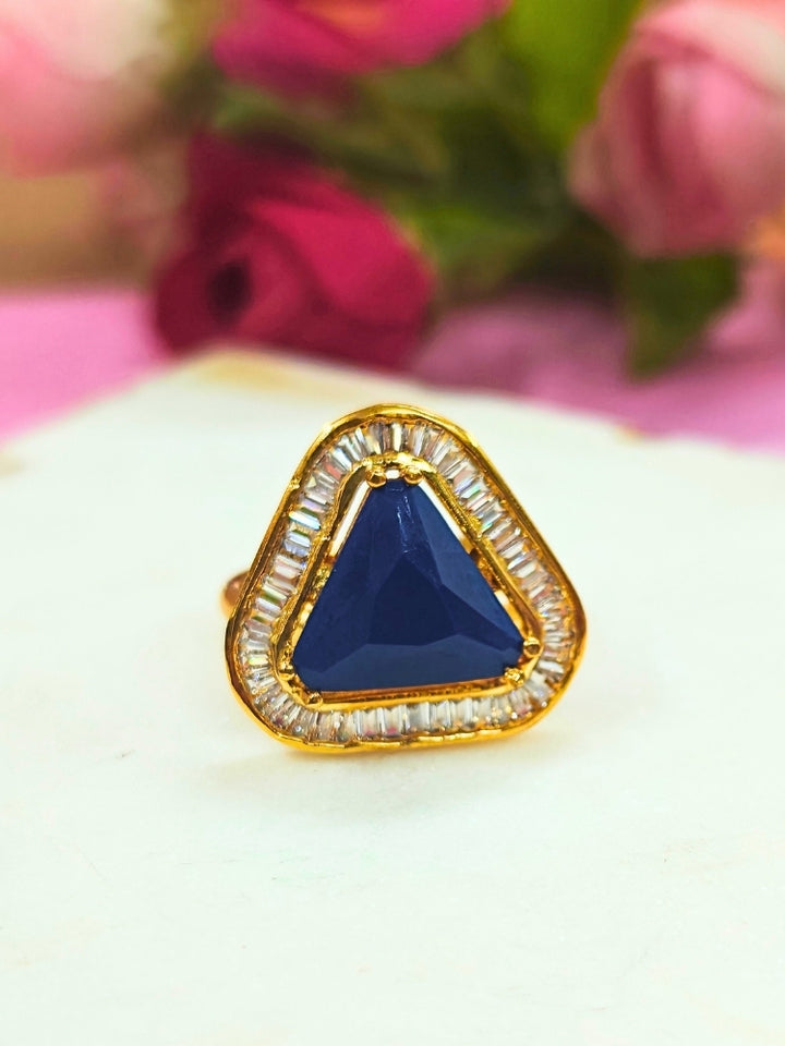 The Penn Blue Ring is an adjustable Blue ring with a triangular Blue gemstone surrounded by clear stones, shown on a white surface with blurred pink and green flowers—ideal for complementing your wedding ceremony dress.