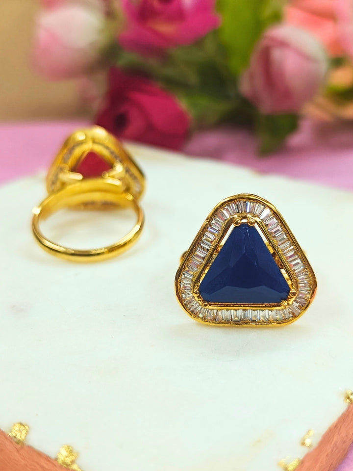 The Penn Blue Ring is an adjustable Blue ring with a triangular Blue gemstone surrounded by clear stones, shown on a white surface with blurred pink and green flowers—ideal for complementing your wedding ceremony dress.