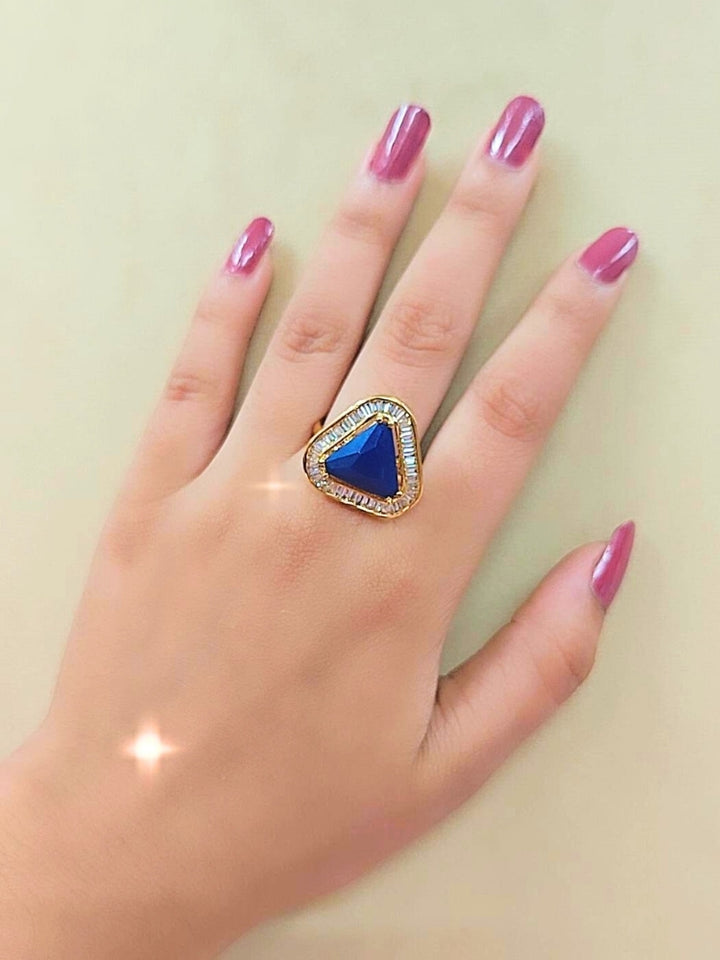 The Penn Blue Ring is an adjustable Blue ring with a triangular Blue gemstone surrounded by clear stones, shown on a white surface with blurred pink and green flowers—ideal for complementing your wedding ceremony dress.