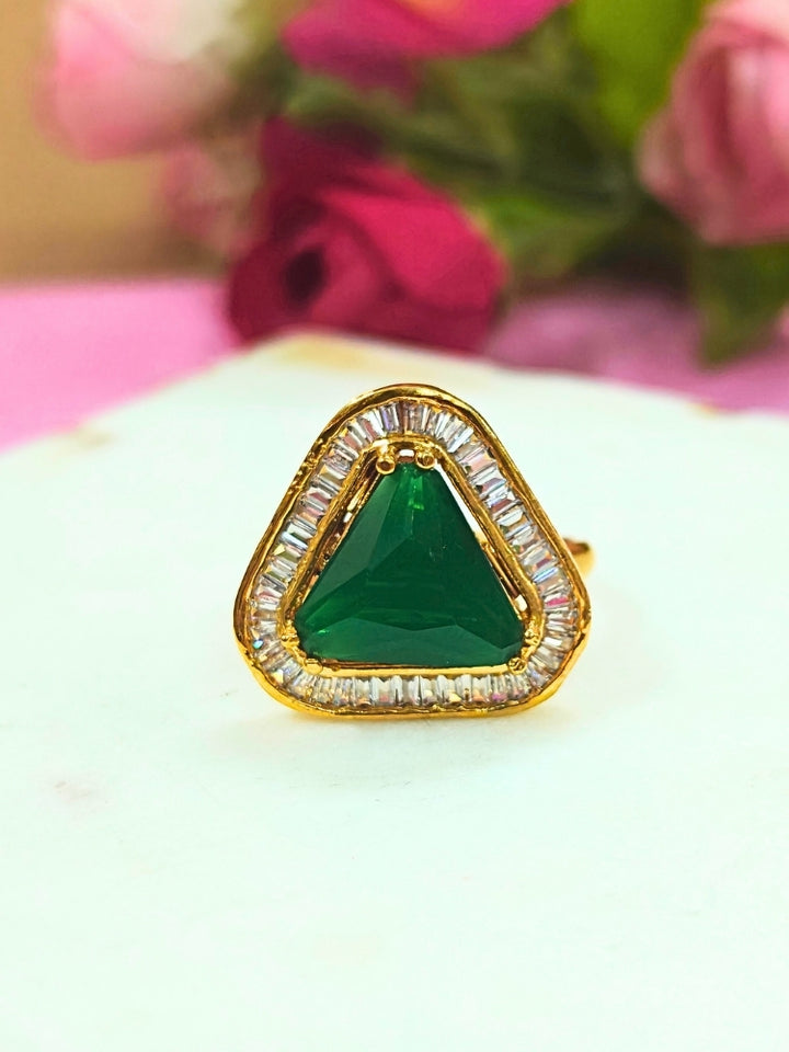 The Penn Green Ring is an adjustable Green ring with a triangular Green gemstone surrounded by clear stones, shown on a white surface with blurred pink and green flowers—ideal for complementing your wedding ceremony dress.