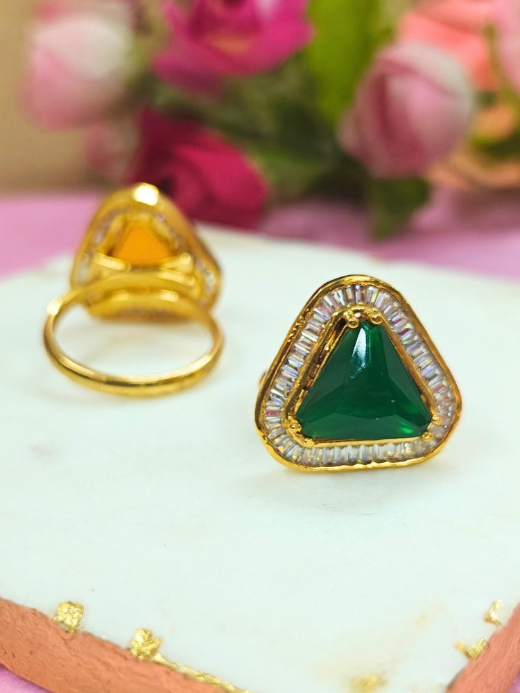 The Penn Green Ring is an adjustable Green ring with a triangular Green gemstone surrounded by clear stones, shown on a white surface with blurred pink and green flowers—ideal for complementing your wedding ceremony dress.