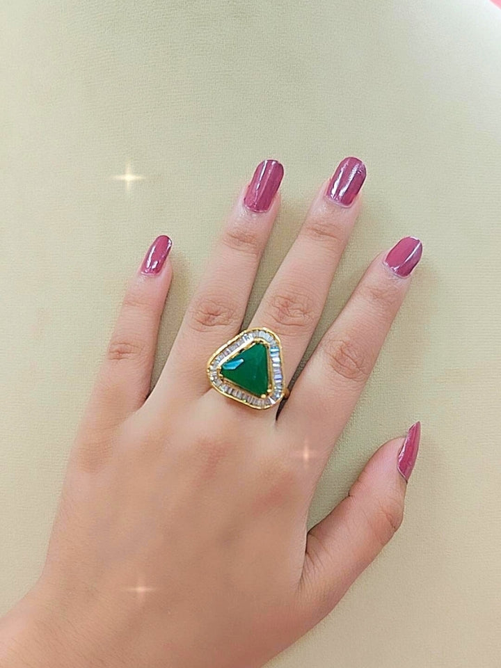The Penn Green Ring is an adjustable Green ring with a triangular Green gemstone surrounded by clear stones, shown on a white surface with blurred pink and green flowers—ideal for complementing your wedding ceremony dress.