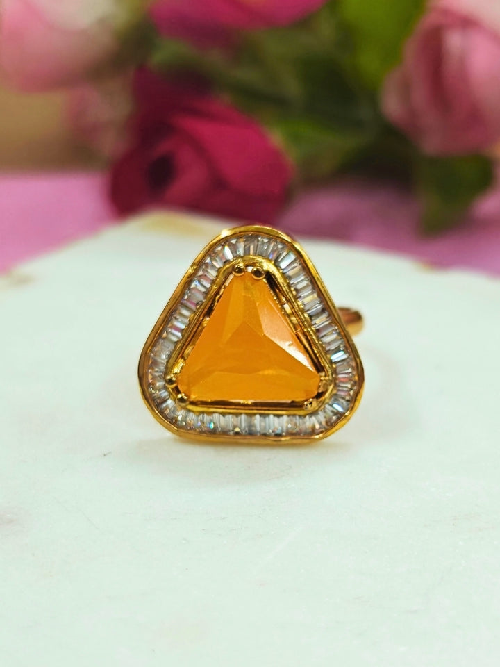 The Penn Yellow Ring is an adjustable yellow ring with a triangular yellow gemstone surrounded by clear stones, shown on a white surface with blurred pink and green flowers—ideal for complementing your wedding ceremony dress.