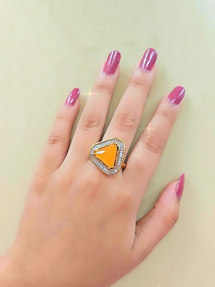 The Penn Yellow Ring is an adjustable Yellow ring with a triangular Yellow gemstone surrounded by clear stones, shown on a white surface with blurred pink and green flowers—ideal for complementing your wedding ceremony dress.