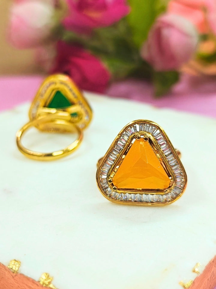 The Penn Yellow Ring is an adjustable Yellow ring with a triangular Yellow gemstone surrounded by clear stones, shown on a white surface with blurred pink and green flowers—ideal for complementing your wedding ceremony dress.