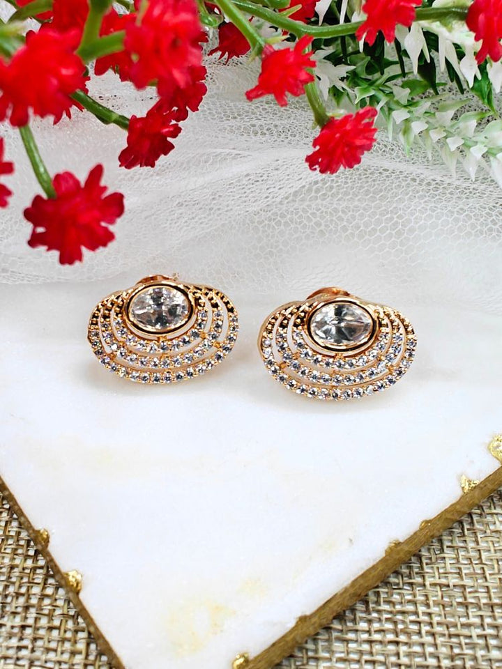 The Pernia stud Earrings are gold-toned, round earrings featuring a large central American Diamond surrounded by rows of smaller sparkling stones, shown on white marble with red flowers and greenery in the background.