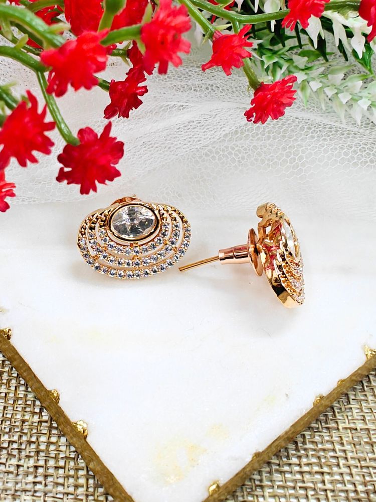 The Pernia stud Earrings are gold-toned, round earrings featuring a large central American Diamond surrounded by rows of smaller sparkling stones, shown on white marble with red flowers and greenery in the background.