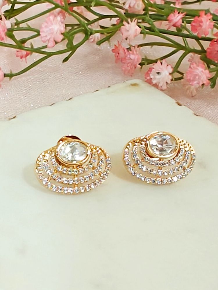 The Pernia stud Earrings are gold-toned, round earrings featuring a large central American Diamond surrounded by rows of smaller sparkling stones, shown on white marble with Pink flowers and greenery in the background.
