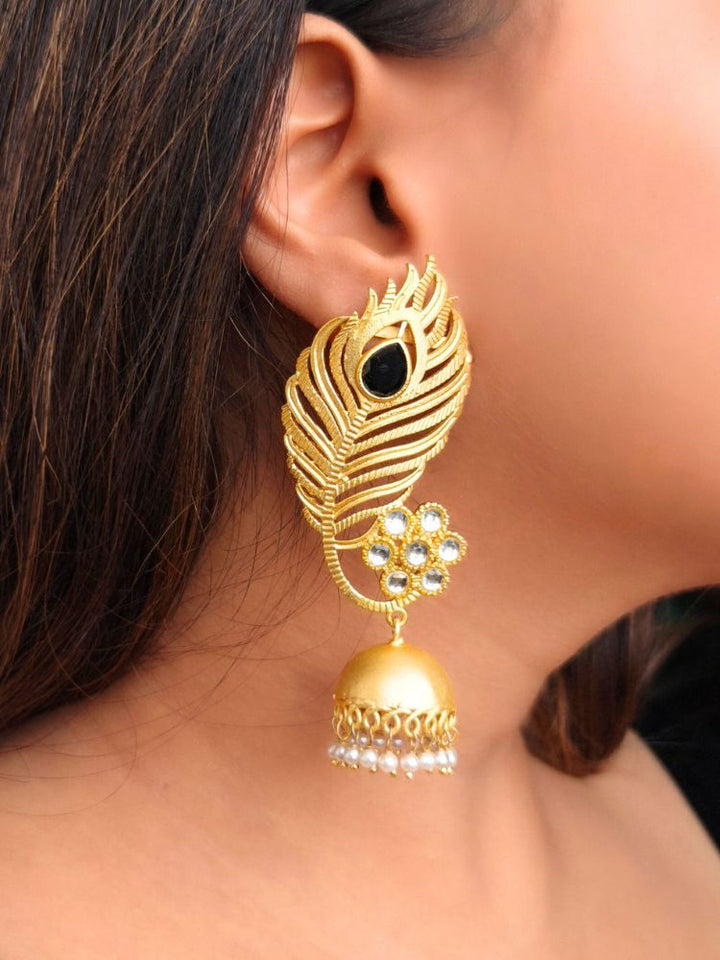 A close-up of a womans ear showcases Pihu Black Earrings—peacock feather-inspired matt gold earrings with Kundan work, a black stone, floral white stones, and a dangling dome featuring pearl accents.