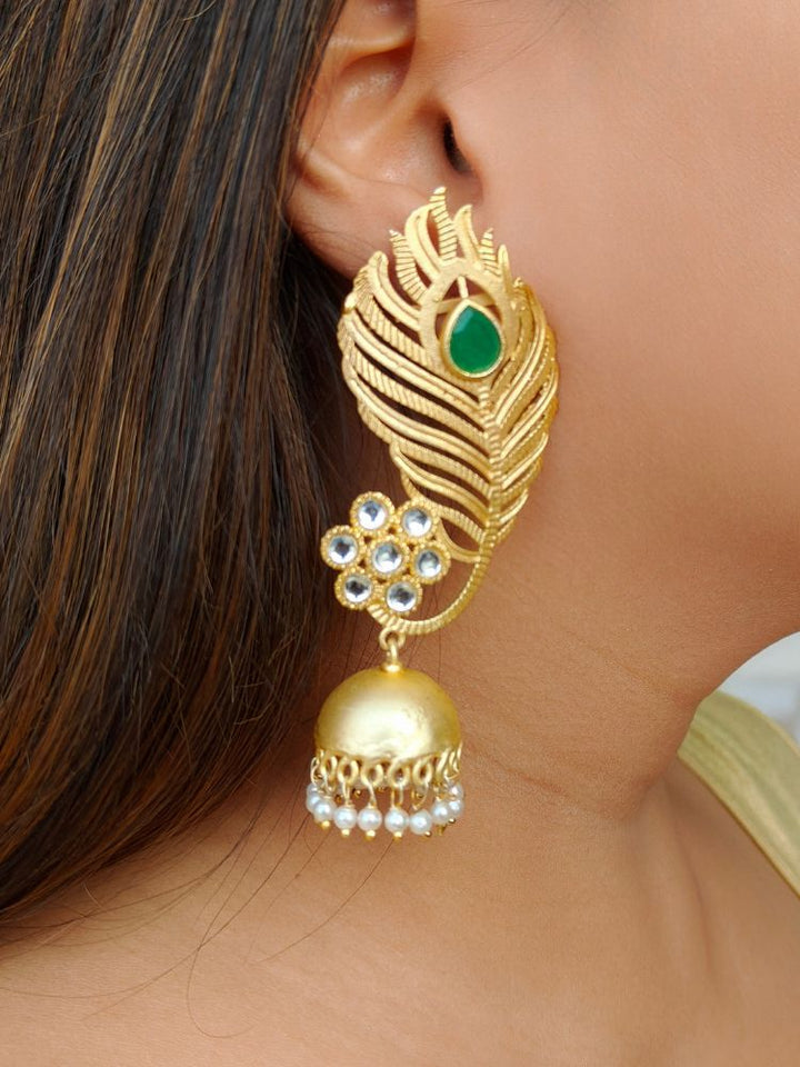A close-up of a womans ear showcases Pihu Green Earrings—peacock feather-inspired matt gold earrings with Kundan work, a Green stone, floral white stones, and a dangling dome featuring pearl accents.