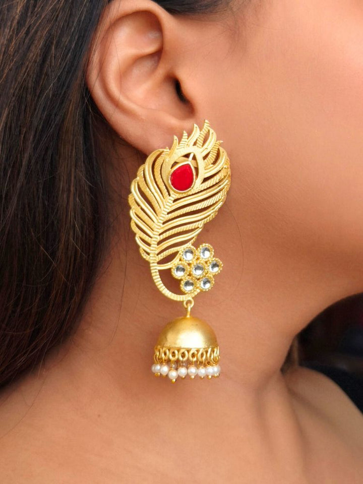 A close-up of a womans ear showcases Pihu Red Earrings—peacock feather-inspired matt gold earrings with Kundan work, a Red stone, floral white stones, and a dangling dome featuring pearl accents.