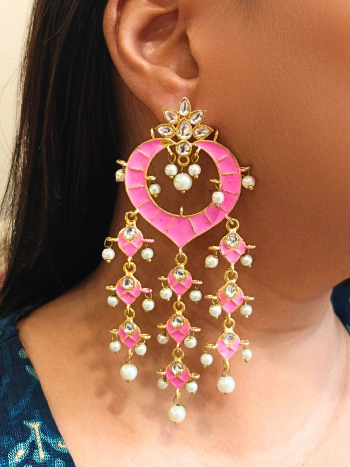 A close-up of a person wearing Pink Ankita Earrings—large, ornate pink and gold earrings with dangling pearls and gemstones in intricate patterns—standing out against dark hair and a blue patterned top. It has meenakari design all over the earrings, and is perfect for wedding functions. 