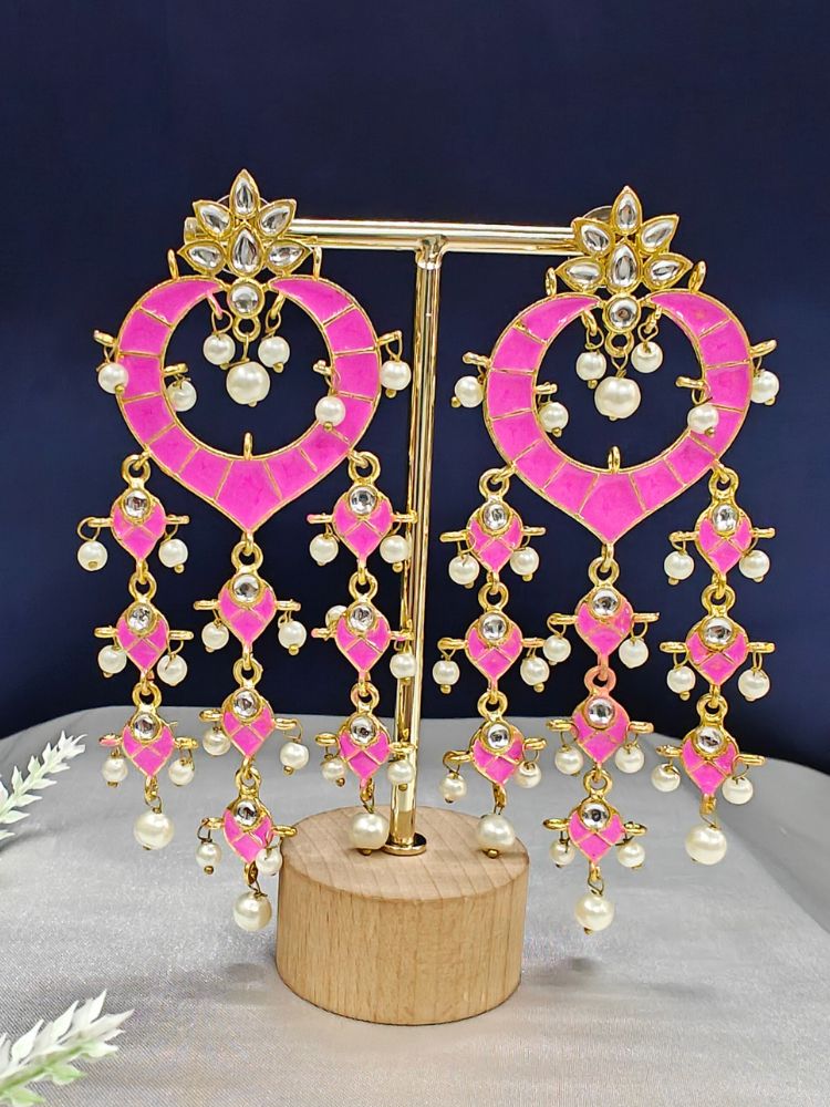 The Pink Ankita Earrings are ornate gold Kundan earrings with pink enamel, intricate Meenakari work, white stones, and dangling pearl beads, shown on a gold stand with a wooden base against a dark background.