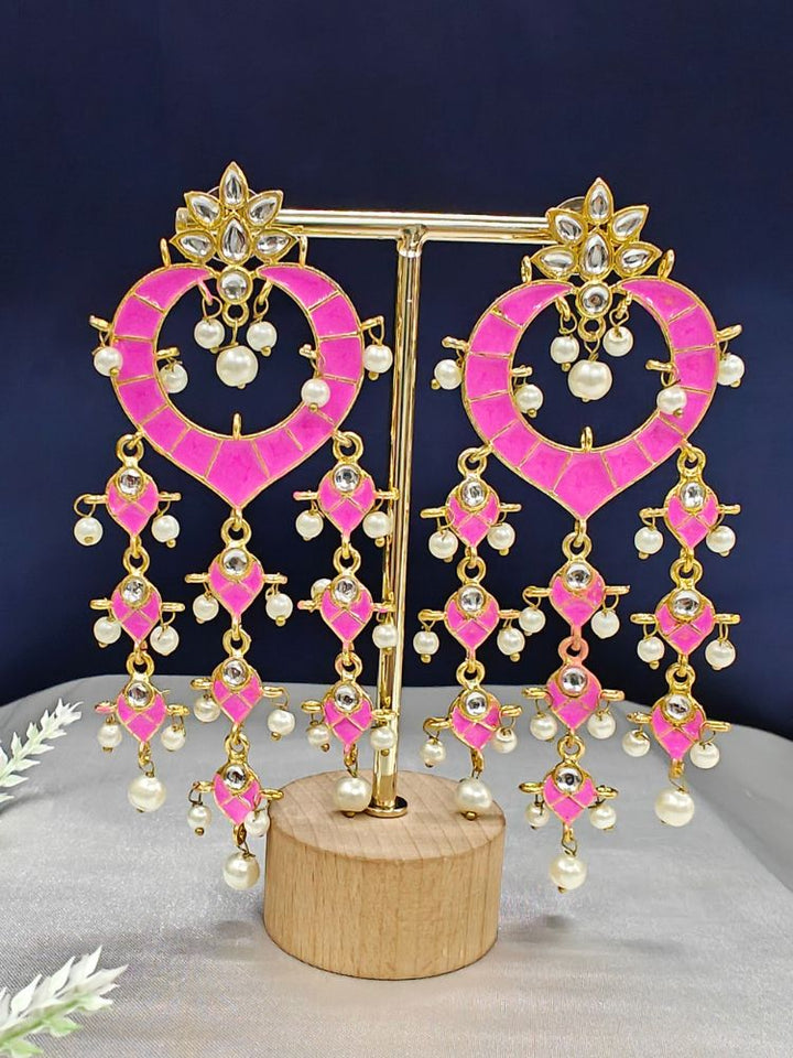 The Pink Ankita Earrings are ornate gold Kundan earrings with pink enamel, intricate Meenakari work, white stones, and dangling pearl beads, shown on a gold stand with a wooden base against a dark background.