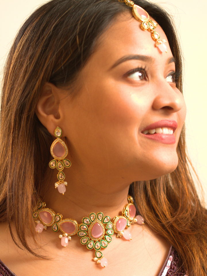 A woman showcases the Pink Aurora Necklace Set, featuring a maang tikka, chandelier earrings, and choker necklace, while dressed in a Purple and gold outfit against a plain background.