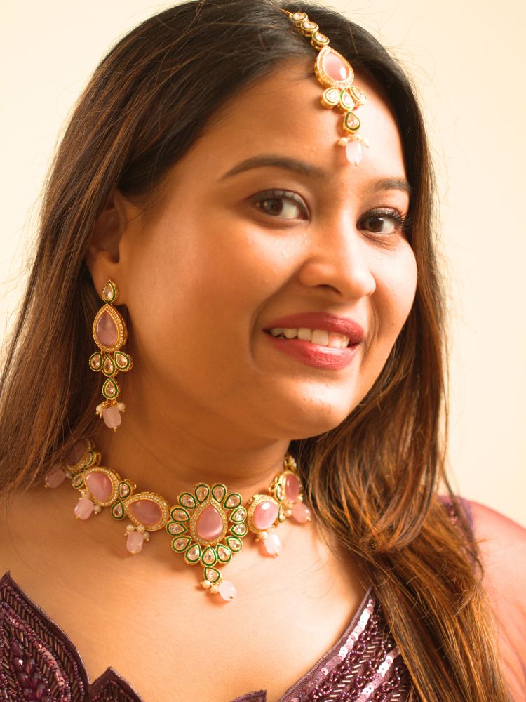 A woman showcases the Pink Aurora Necklace Set, featuring a maang tikka, chandelier earrings, and choker necklace, while dressed in a Purple and gold outfit against a plain background.