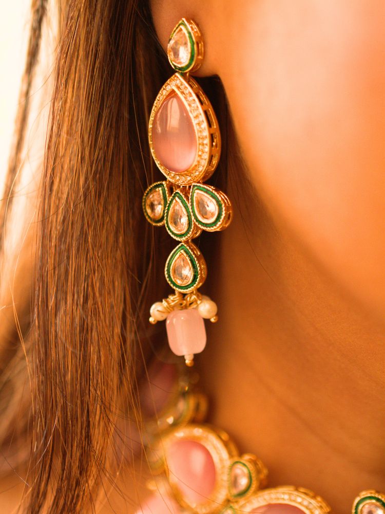 A woman showcases the Pink Aurora Necklace Set, featuring a maang tikka, chandelier earrings, and choker necklace, while dressed in a Purple and gold outfit against a plain background.