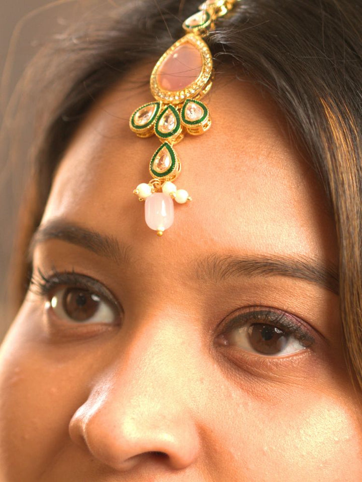 A woman showcases the Pink Aurora Necklace Set, featuring a maang tikka, chandelier earrings, and choker necklace, while dressed in a Purple and gold outfit against a plain background.