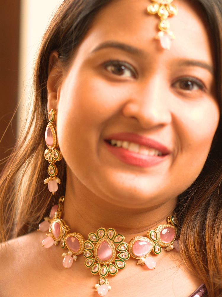 A woman showcases the Pink Aurora Necklace Set, featuring a maang tikka, chandelier earrings, and choker necklace, while dressed in a Purple and gold outfit against a plain background.