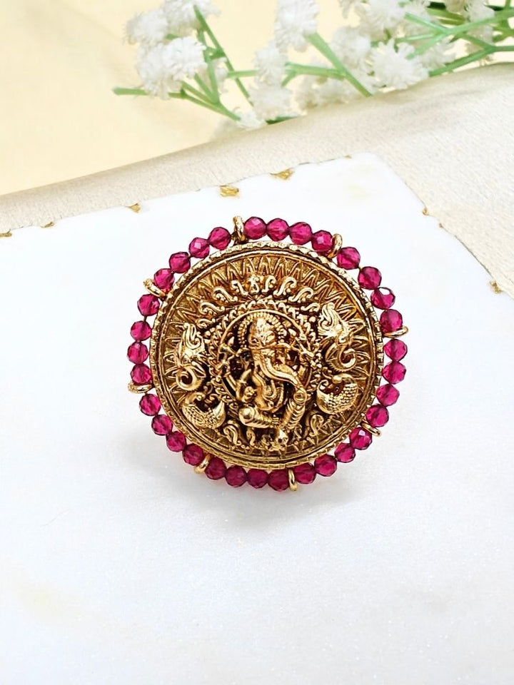 The Pink Bheema Ring is a round, gold-toned adjustable ring featuring Ganeshji made in intricate detailing at the center, encircled by Pink crystal stones. Displayed on white cloth with small flowers, its ideal for enhancing your wedding ceremony outfit.