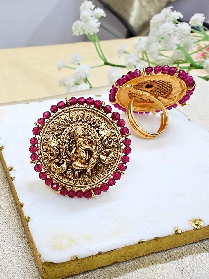 The Pink Bheema Ring is a round, gold-toned adjustable ring featuring Ganeshji made in intricate detailing at the center, encircled by Pink crystal stones. Displayed on white cloth with small flowers, its ideal for enhancing your wedding ceremony outfit.