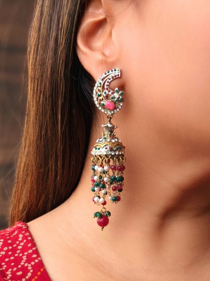 A close-up of a woman with straight brown hair wearing the Pink Charu Earrings—large, ornate Victorian-style earrings adorned with intricate designs, colorful beads, dangling elements, and striking red, green, and gold accents.
