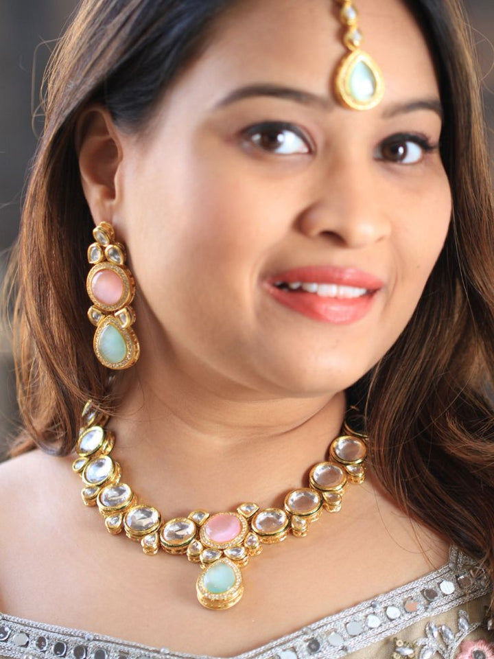 A woman wearing a silver outfit and long brown hair smiles at the camera, showcasing the Pink & Green Dhara Kundan Necklace Set—gold jewelry with pink, green, and clear Monalisa stones—complete with necklace, earrings, and maang tikka.