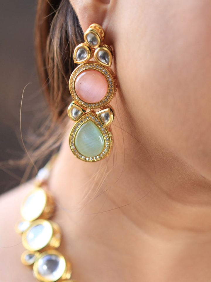 A woman wearing a silver outfit and long brown hair smiles at the camera, showcasing the Pink & Green Dhara Kundan Necklace Set—gold jewelry with pink, green, and clear Monalisa stones—complete with necklace, earrings, and maang tikka.