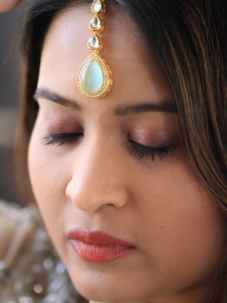 A woman wearing a silver outfit and long brown hair smiles at the camera, showcasing the Pink & Green Dhara Kundan Necklace Set—gold jewelry with pink, green, and clear Monalisa stones—complete with necklace, earrings, and maang tikka.