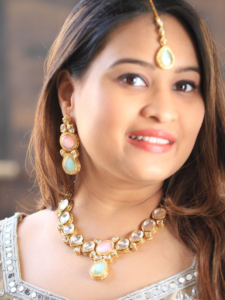A woman wearing a silver outfit and long brown hair smiles at the camera, showcasing the Pink & Green Dhara Kundan Necklace Set—gold jewelry with pink, green, and clear Monalisa stones—complete with necklace, earrings, and maang tikka.