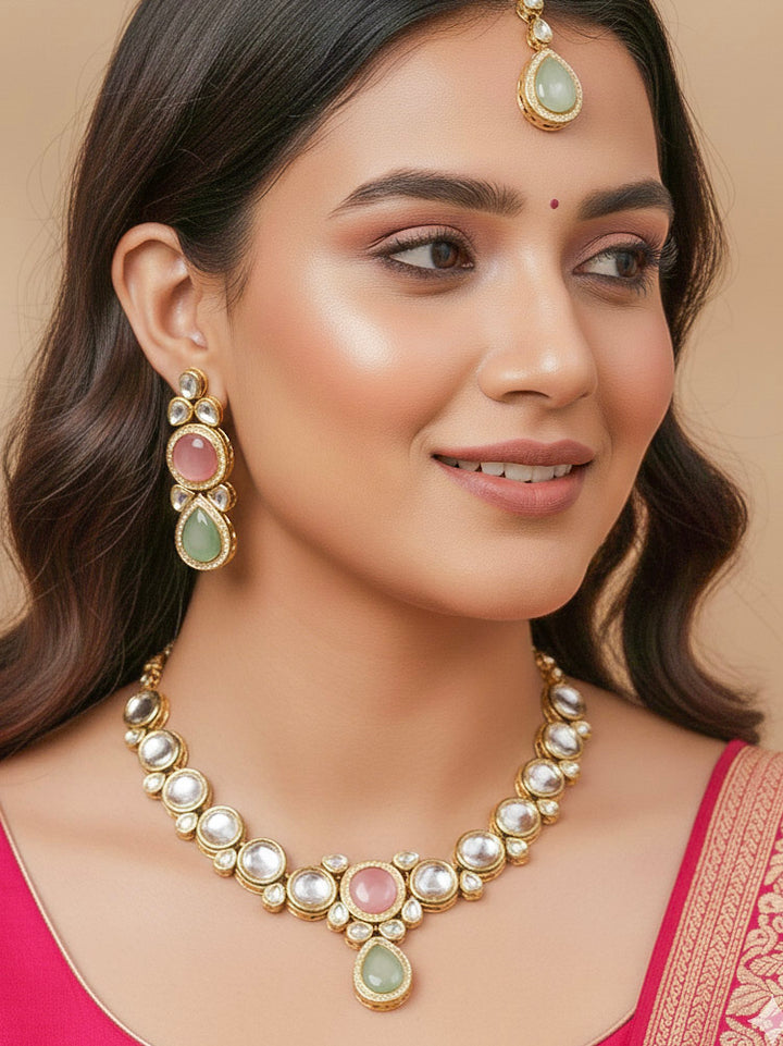 A woman wearing a silver outfit and long brown hair smiles at the camera, showcasing the Pink & Green Dhara Kundan Necklace Set—gold jewelry with pink, green, and clear Monalisa stones—complete with necklace, earrings, and maang tikka.
