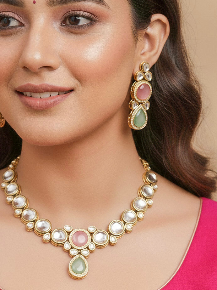 A woman wearing a silver outfit and long brown hair smiles at the camera, showcasing the Pink & Green Dhara Kundan Necklace Set—gold jewelry with pink, green, and clear Monalisa stones—complete with necklace, earrings, and maang tikka.
