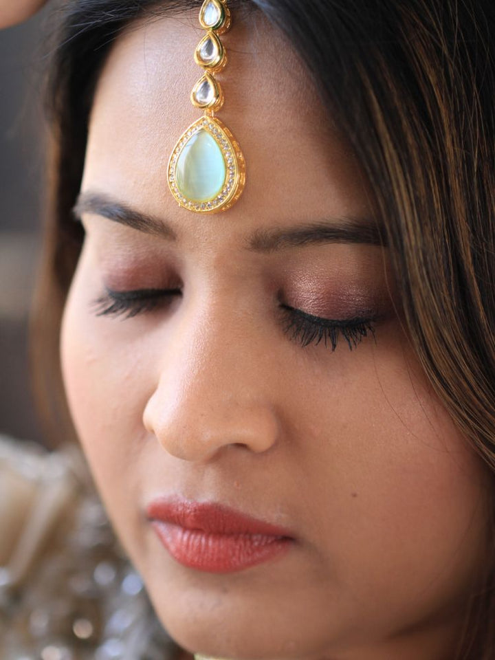 A woman wearing a silver outfit and long brown hair smiles at the camera, showcasing the Pink & Green Dhara Kundan Necklace Set—gold jewelry with pink, green, and clear Monalisa stones—complete with necklace, earrings, and maang tikka.
