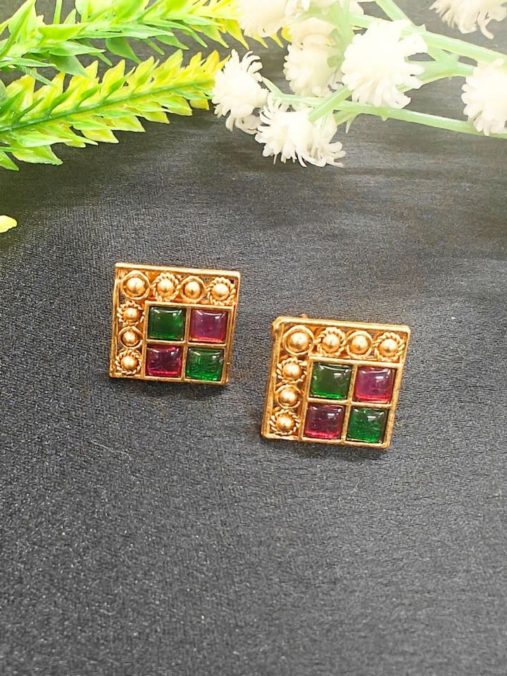 The Pink & Green Vanita Earrings are square gold earrings with Two Green & Pink gemstones and gold bead detailing, shown on a black surface with yellow and white flowers—ideal for adding traditional flair to your Kurti ensemble.