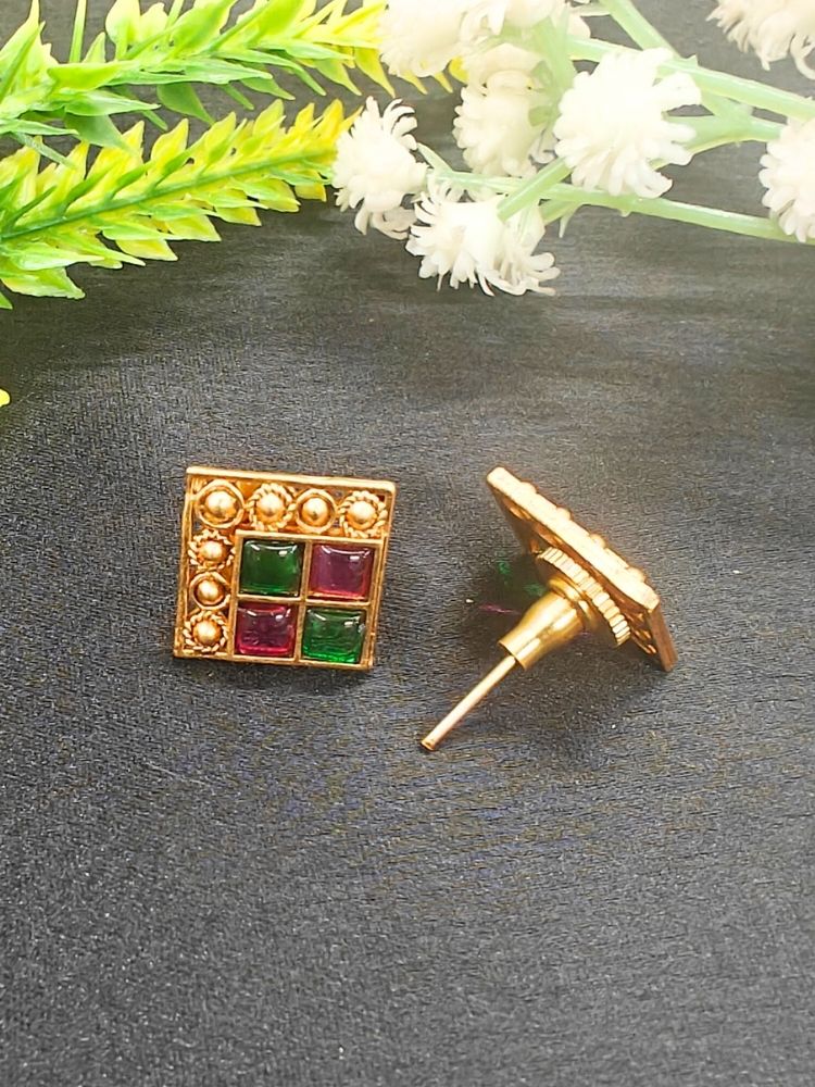 The Pink & Green Vanita Earrings are square gold earrings with Two Green & Pink gemstones and gold bead detailing, shown on a black surface with yellow and white flowers—ideal for adding traditional flair to your Kurti ensemble.