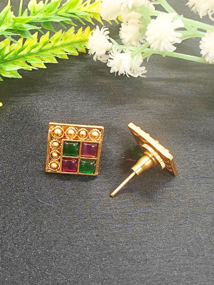 The Pink & Green Vanita Earrings are square gold earrings with Two Green & Pink gemstones and gold bead detailing, shown on a black surface with yellow and white flowers—ideal for adding traditional flair to your Kurti ensemble.