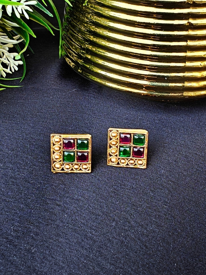 The Pink & Green Vanita Earrings are square gold earrings with Two Green & Pink gemstones and gold bead detailing, shown on a black surface with yellow and white flowers—ideal for adding traditional flair to your Kurti ensemble.