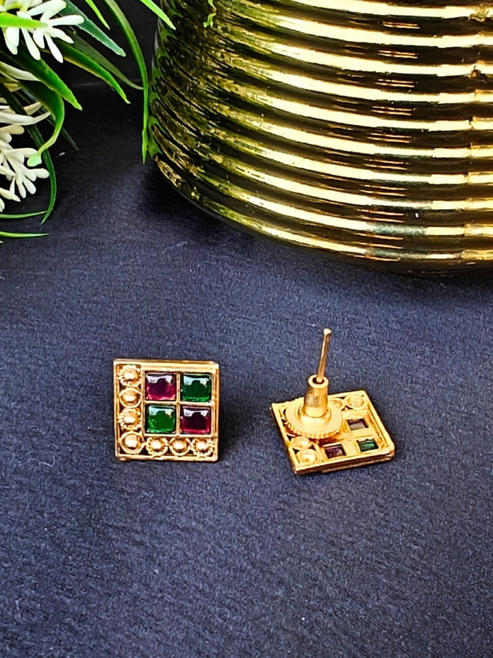 The Pink & Green Vanita Earrings are square gold earrings with Two Green & Pink gemstones and gold bead detailing, shown on a black surface with yellow and white flowers—ideal for adding traditional flair to your Kurti ensemble.