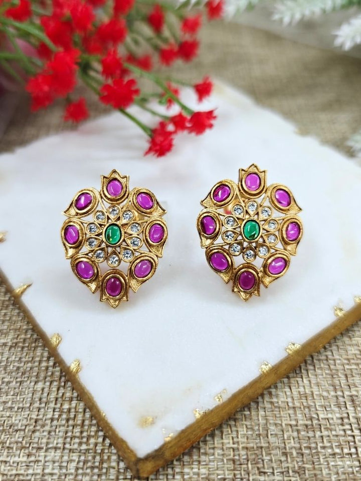 The Pink Mohini stud Earrings are ornate gold earrings embellished with American Diamonds and vibrant gemstones in Pink, shown on a white marble slab with red flowers in the background.