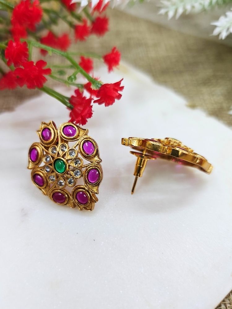The Pink Mohini stud Earrings are ornate gold earrings embellished with American Diamonds and vibrant gemstones in Pink, shown on a white marble slab with red flowers in the background.