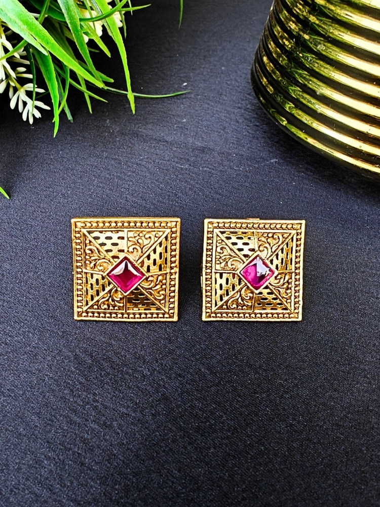 The Pink Mudita stud Earrings feature square gold filigree with intricate patterns and a red gemstone at the center, elegantly displayed on a black surface beside green leaves and a gold-striped container.