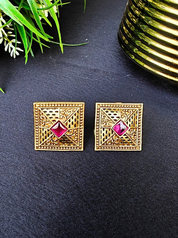 The Pink Mudita stud Earrings feature square gold filigree with intricate patterns and a red gemstone at the center, elegantly displayed on a black surface beside green leaves and a gold-striped container.