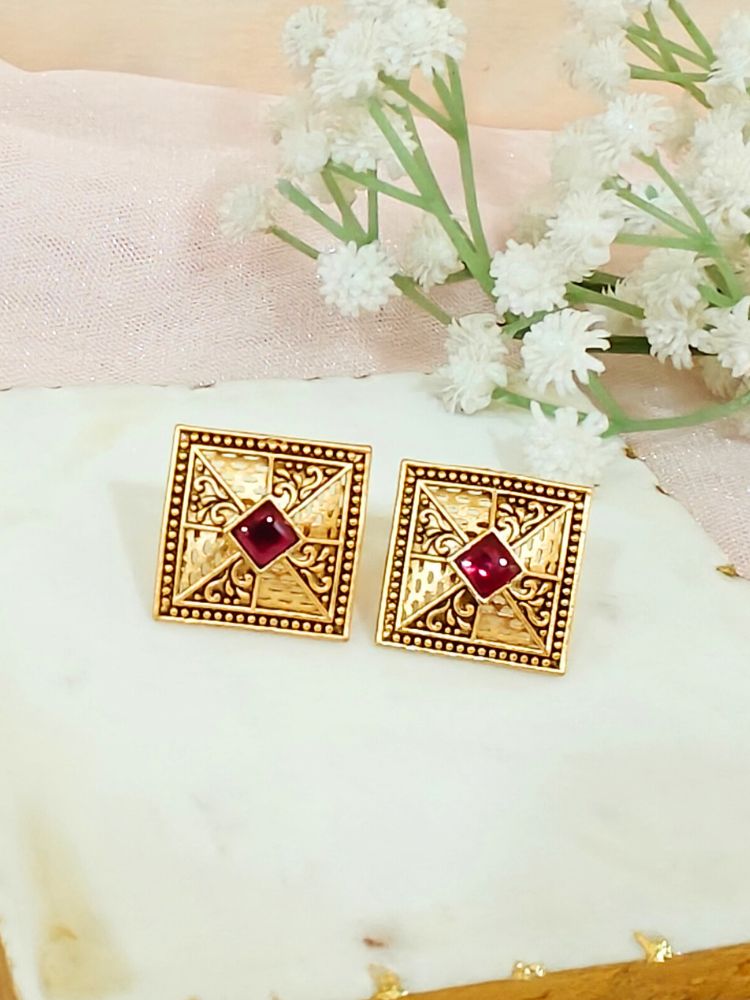 The Pink Mudita stud Earrings feature square gold filigree with intricate patterns and a red gemstone at the center, elegantly displayed on a White Marble surface beside White Flower and a gold-striped container.